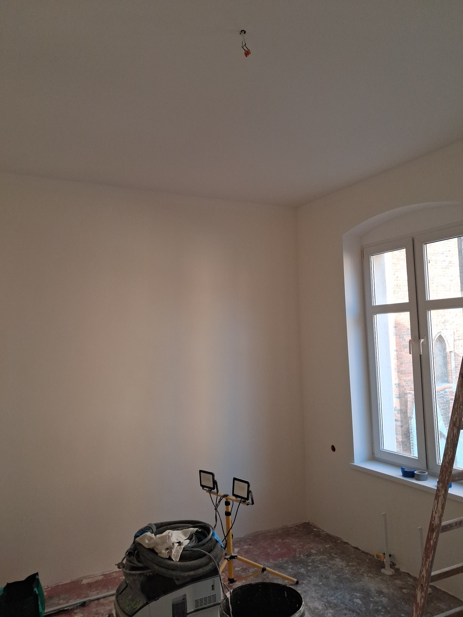 Wnętrze pokoju po świeżym malowaniu ścian na jasny odcień. Widoczne okno z białymi ramami, drabina oparta o ścianę, lampa budowlana i sprzęt malarski.