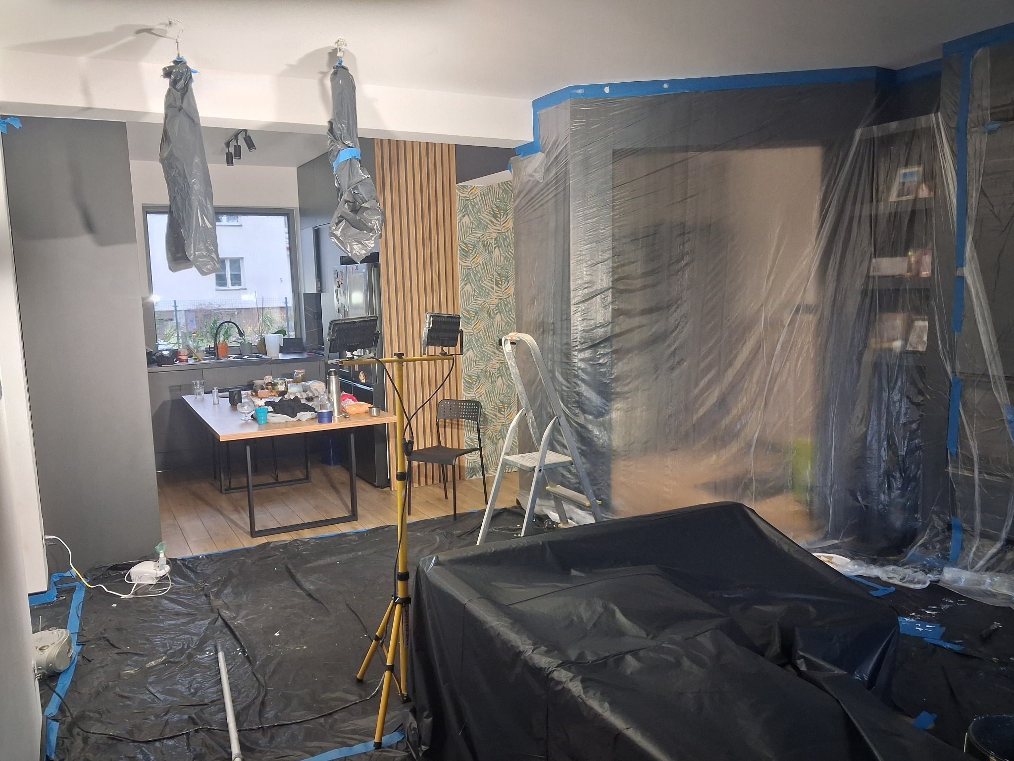 Przygotowanie do prac wykończeniowych: pomieszczenie zabezpieczone folią malarską, widoczna drabina i lampa na statywie. Elementy wyposażenia zakryte.