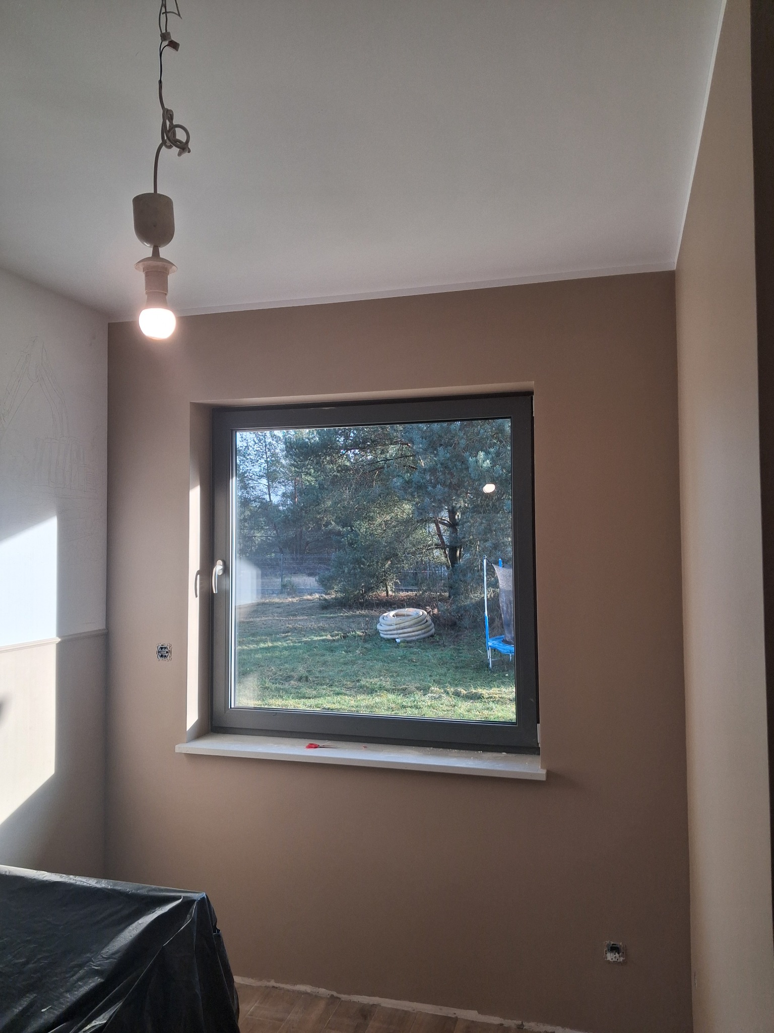 Pokój po malowaniu ścian na beżowo, widoczne okno z widokiem na zewnątrz, sufit pomalowany na biało, na podłodze panele. W rogu pokoju widoczna zakryta folią podłoga.