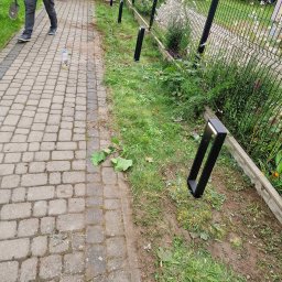 HusariiVolt - Czarna, nowoczesna lampa ogrodowa typu słupek oświetla ścieżkę z kostki brukowej. W tle siatka ogrodzeniowa i zieleń. Widoczna butelka i fragment osoby.