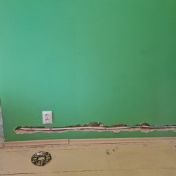 HusariiVolt - Zielona ściana z gniazdkiem i bruzdą, w której widoczne są białe kable elektryczne. Na podłodze leży czarna osłona kabla i widać fragment osoby z wiertarką.