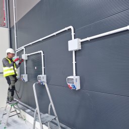 HusariiVolt - Elektryk na drabinie montuje białe kable i gniazda na szarej, żebrowanej ścianie w hali. Widoczne narzędzia i elementy instalacji. Praca w toku.