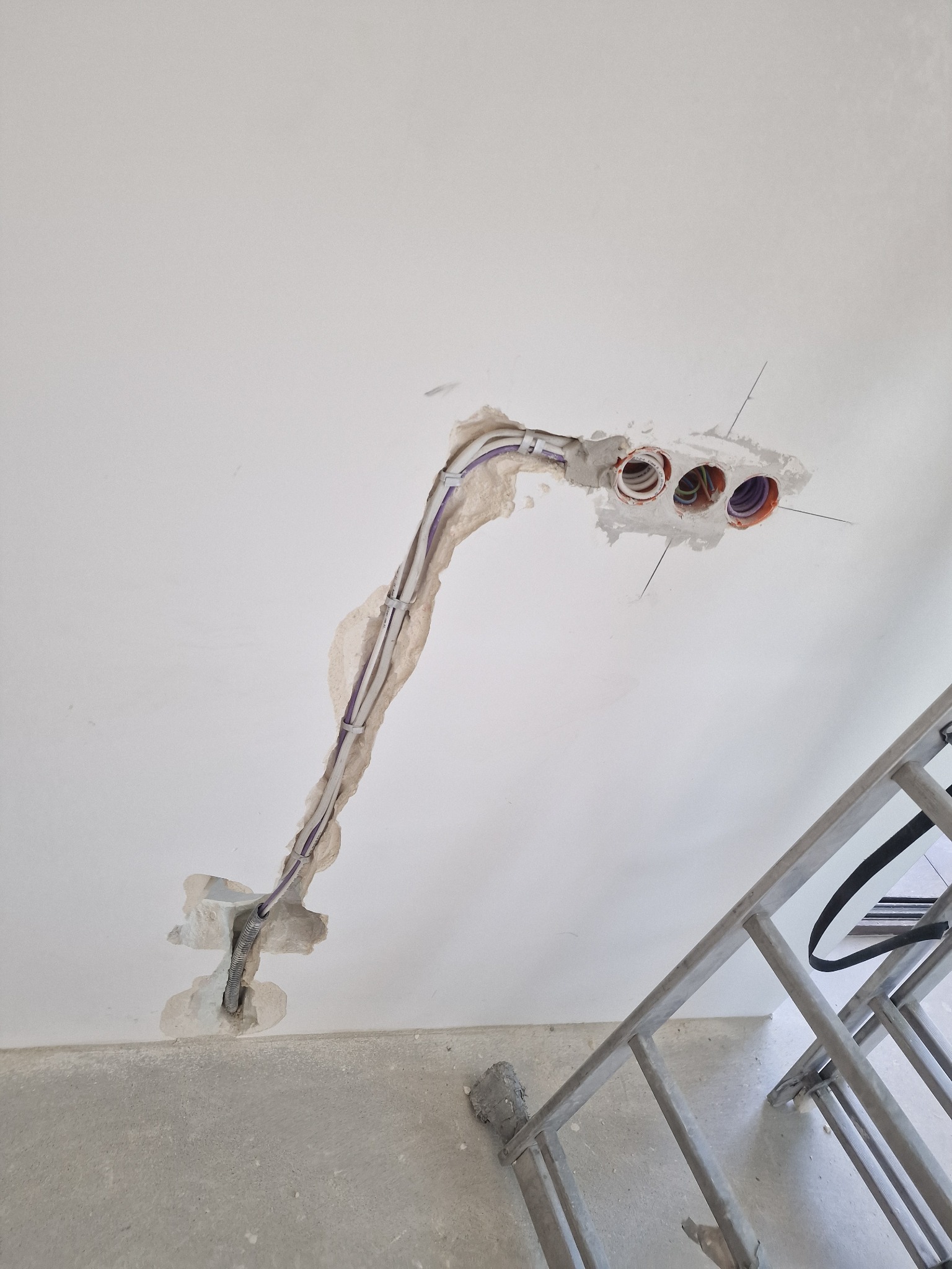 Pionowy kadr: przewody elektryczne w ścianie, prowadzone do trzech otworów z pomarańczowymi rurkami. Widoczny fragment drabiny malarskiej. Prace w trakcie.
