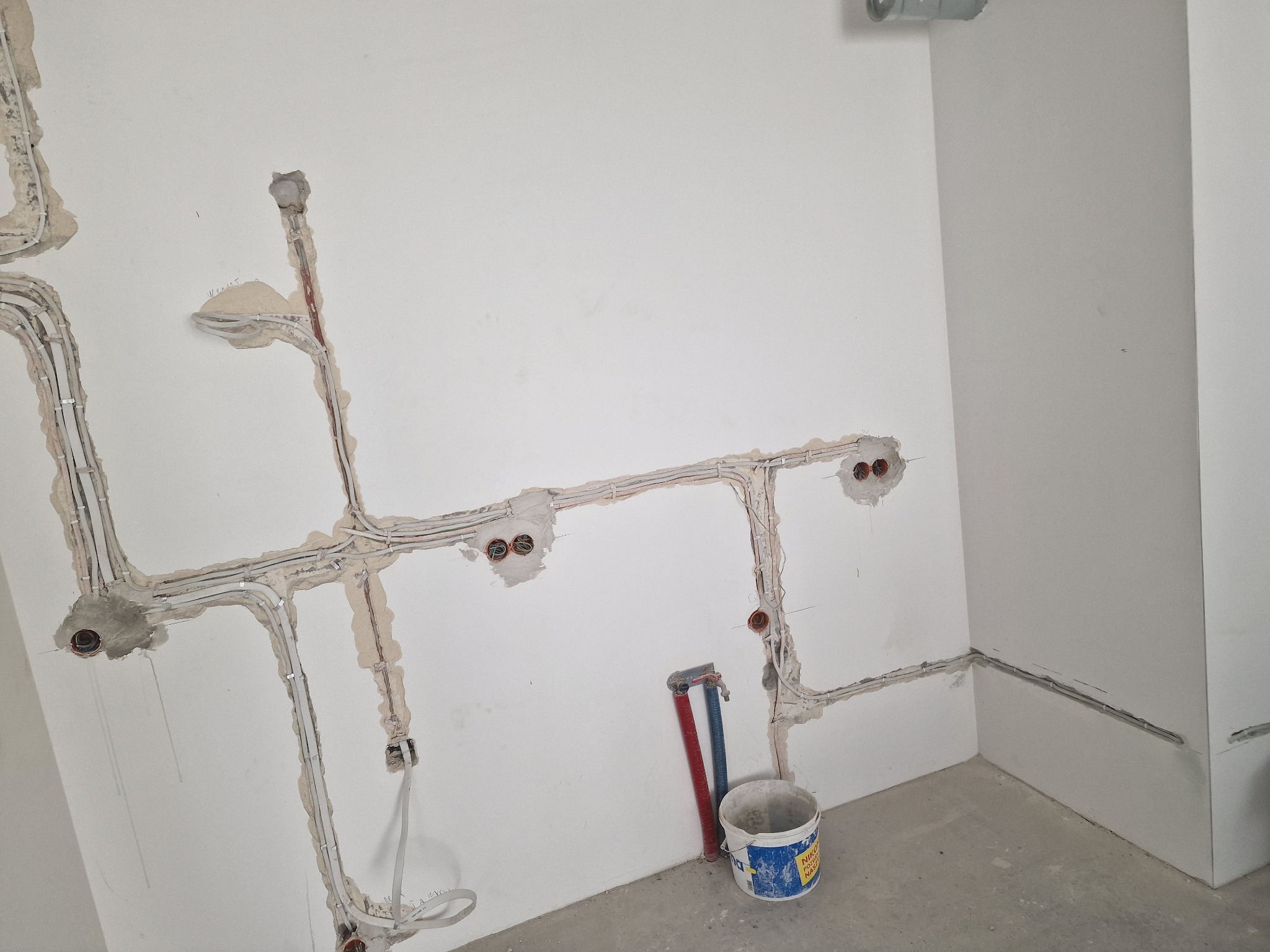 Wykute w ścianie kable elektryczne, widoczne puszki instalacyjne, fragment rur i wiadro. Prace instalacyjne w surowym wnętrzu, przygotowane do dalszego etapu wykończenia.