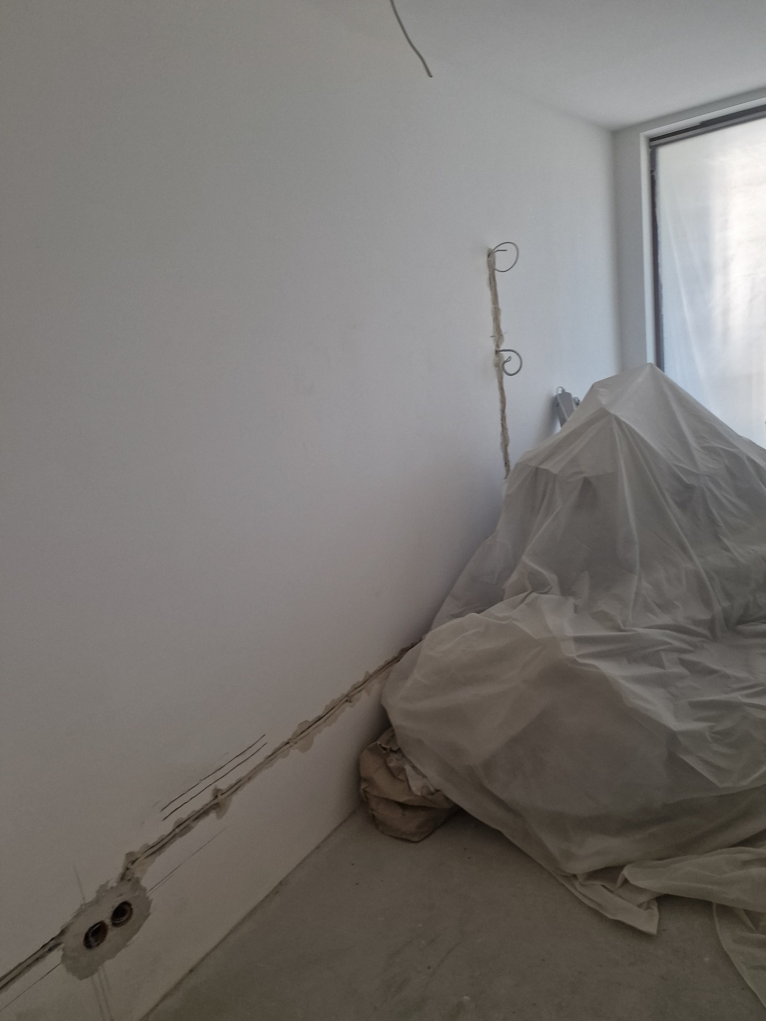 Biała ściana z wyżłobieniami na kable elektryczne i wystającymi przewodami, zakryte meble folią malarską. W tle okno z zasłoniętą szybą.