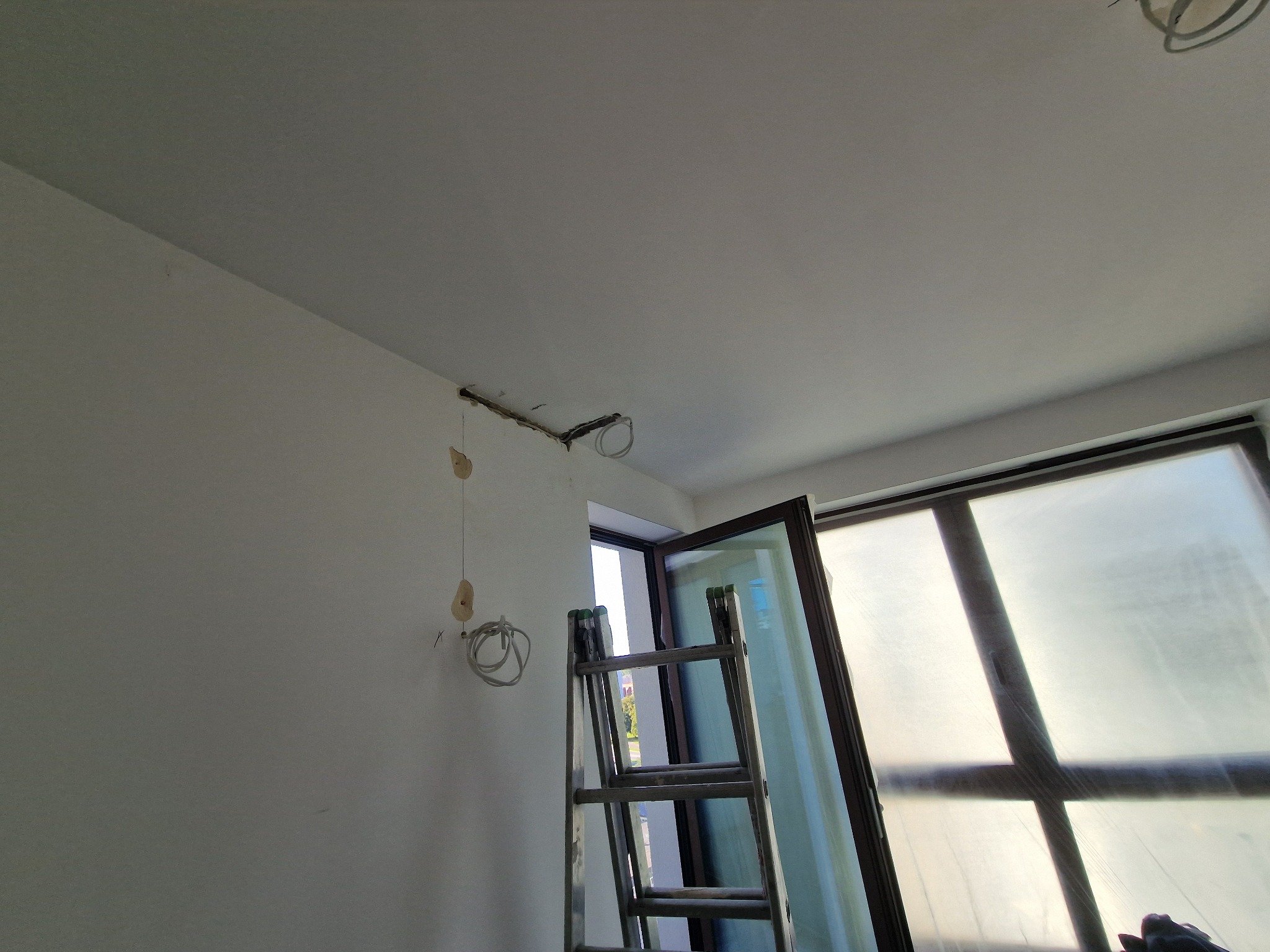 Przygotowanie do montażu oświetlenia sufitowego: widoczne przewody i puszki instalacyjne na białej ścianie, drabina oparta o okno zabezpieczone folią.