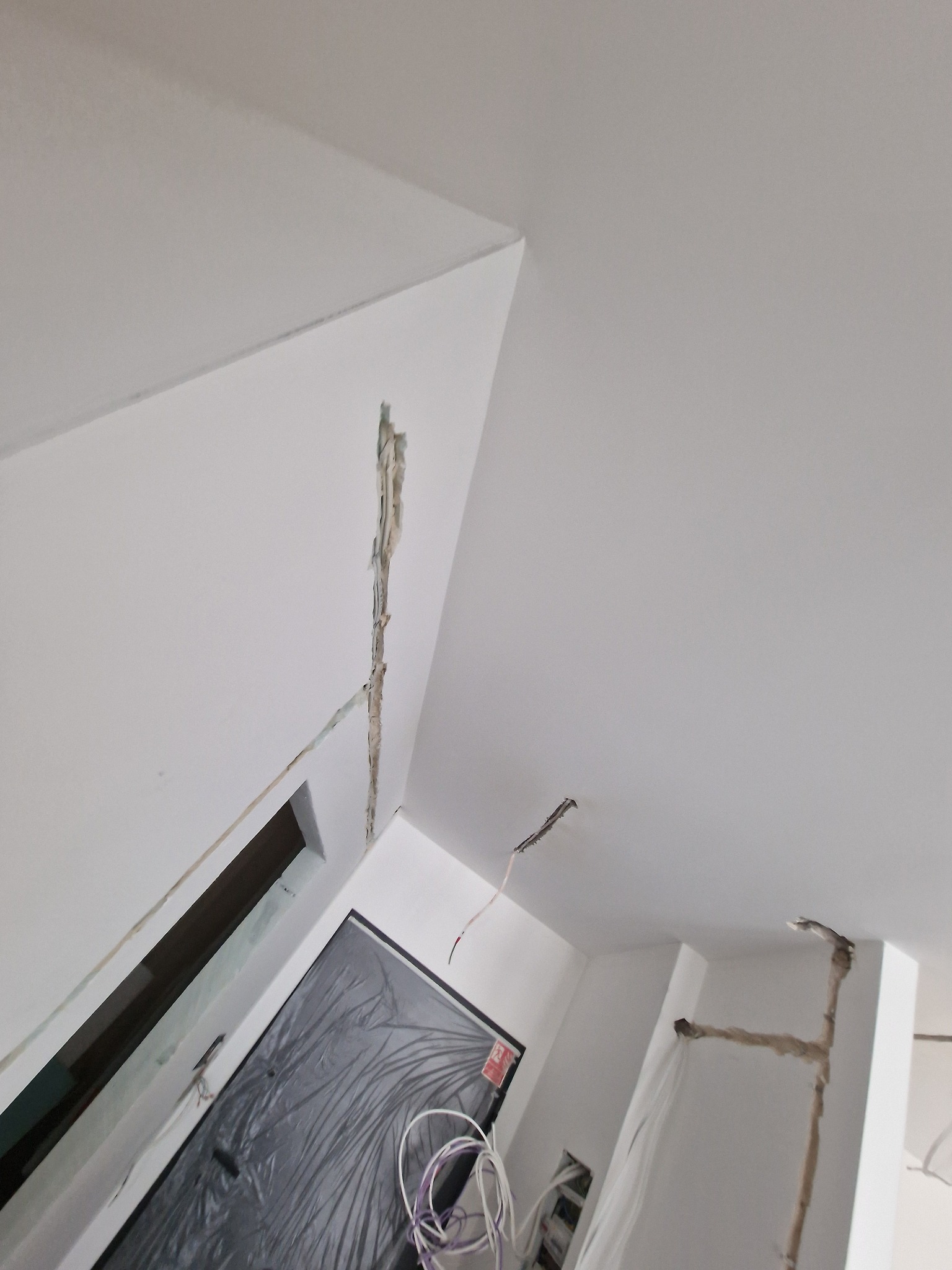 Białe ściany w trakcie prac remontowych. Widoczne kable elektryczne wystające ze ścian i sufitu. Otwór w ścianie zakryty folią. Ujęcie z dołu, pod kątem.