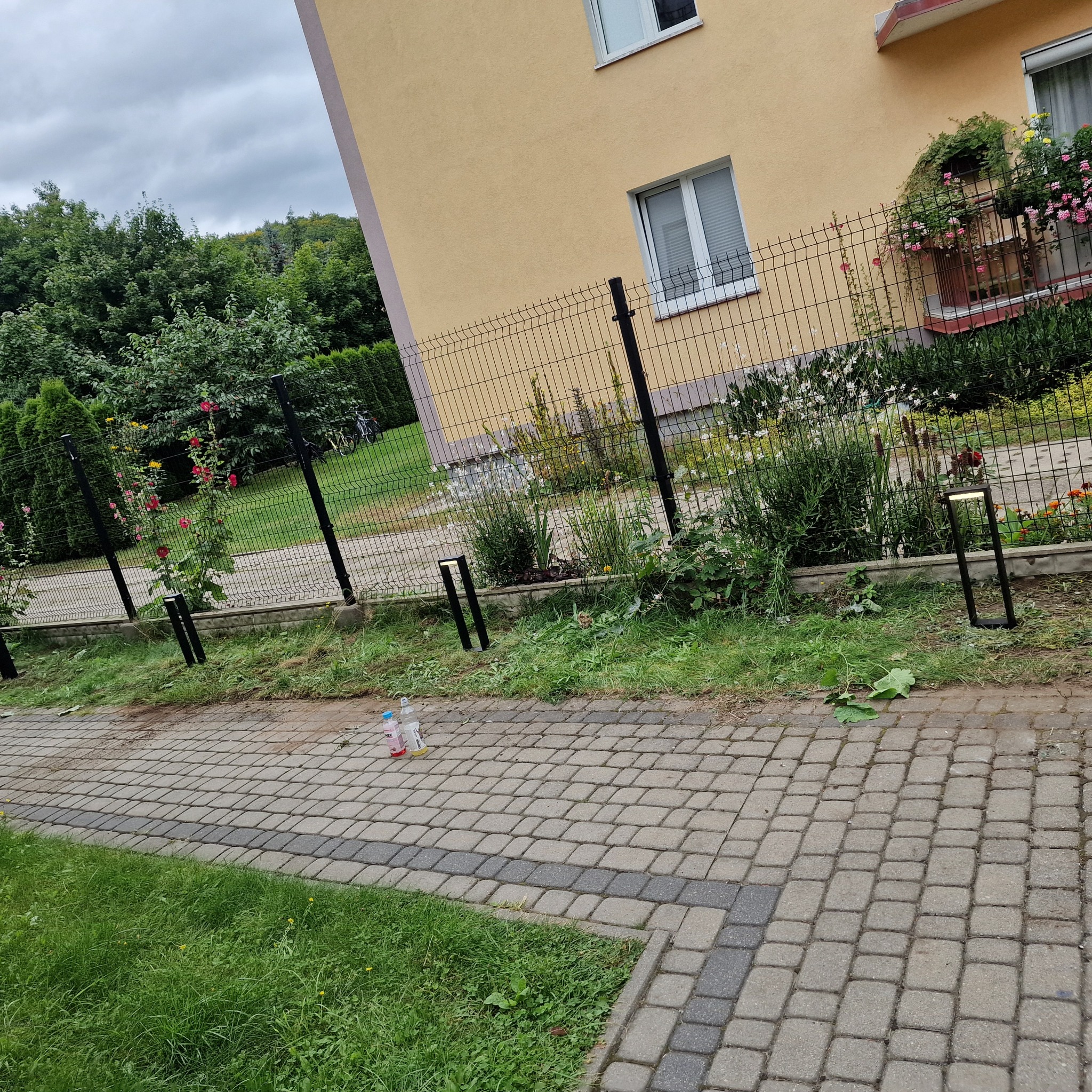 Metalowe ogrodzenie panelowe z lampami ogrodowymi wzdłuż ścieżki z kostki brukowej przed budynkiem z balkonem.