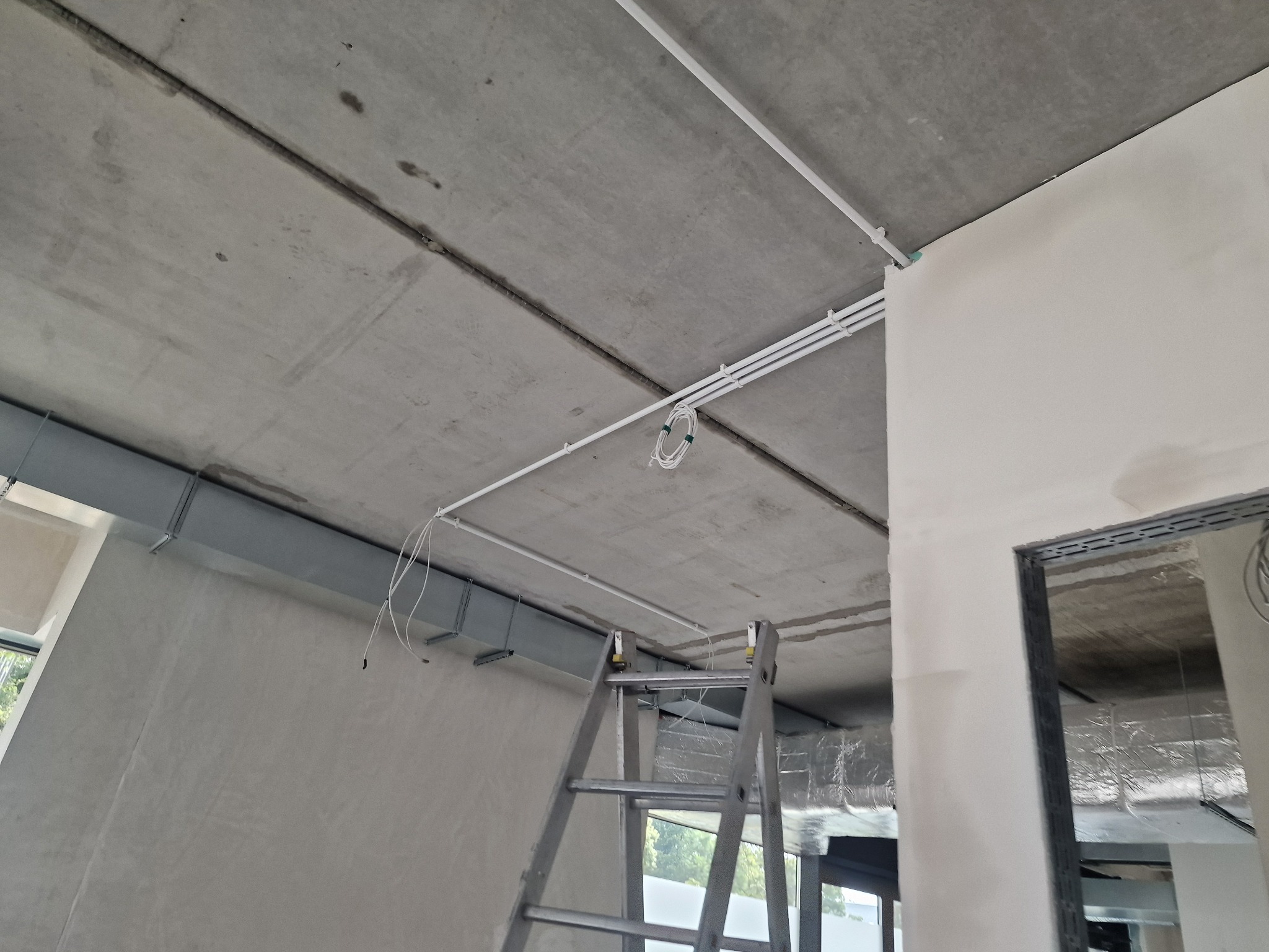 Montaż białych rurek instalacji elektrycznej pod betonowym sufitem. Widoczna drabina i przewody. Surowy stan wykończenia wnętrza.
