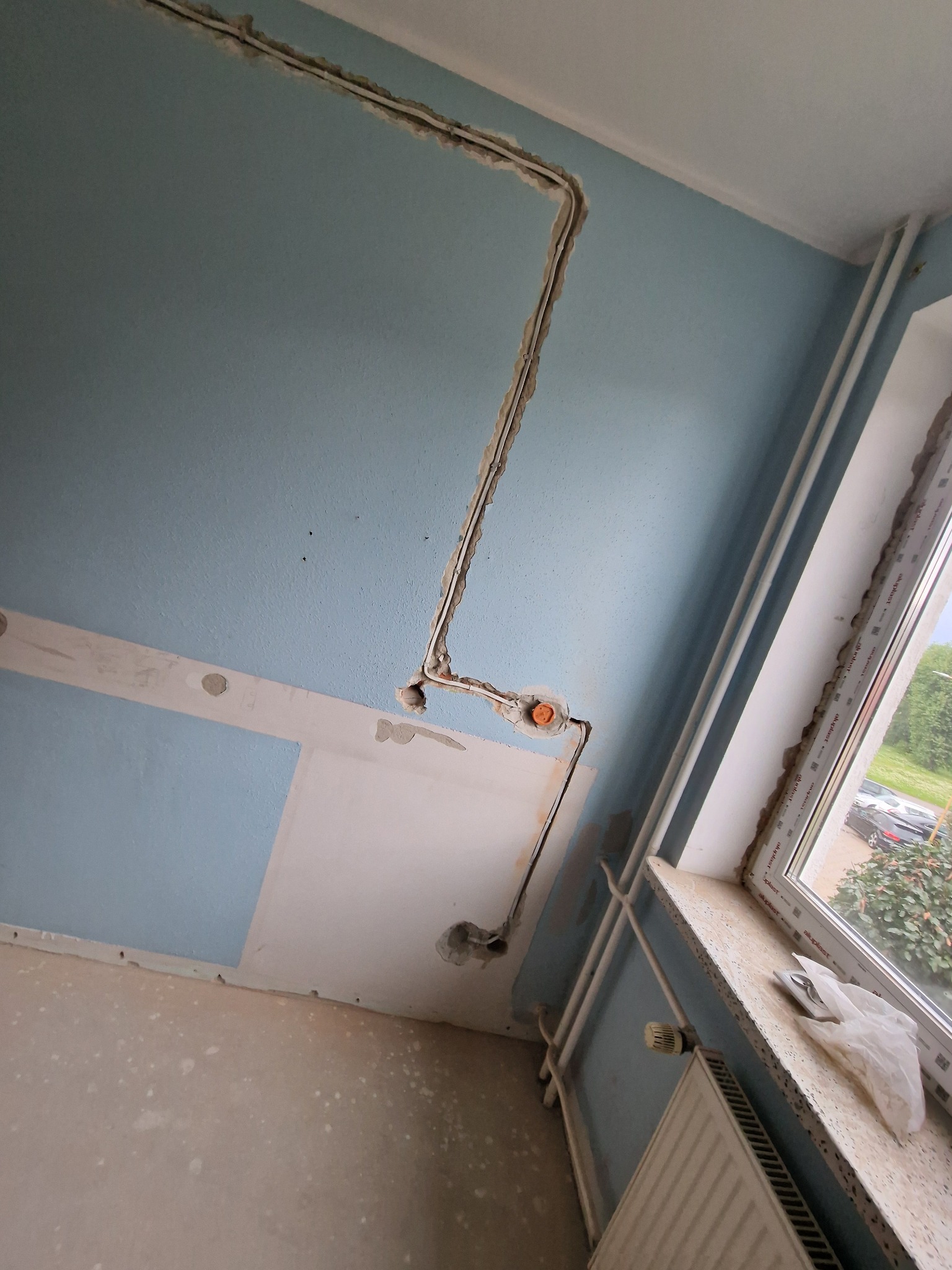 Ściana z wyfrezowanymi bruzdami pod instalację elektryczną, widoczne puszki instalacyjne. Częściowo położona gładź, okno z grzejnikiem w tle.