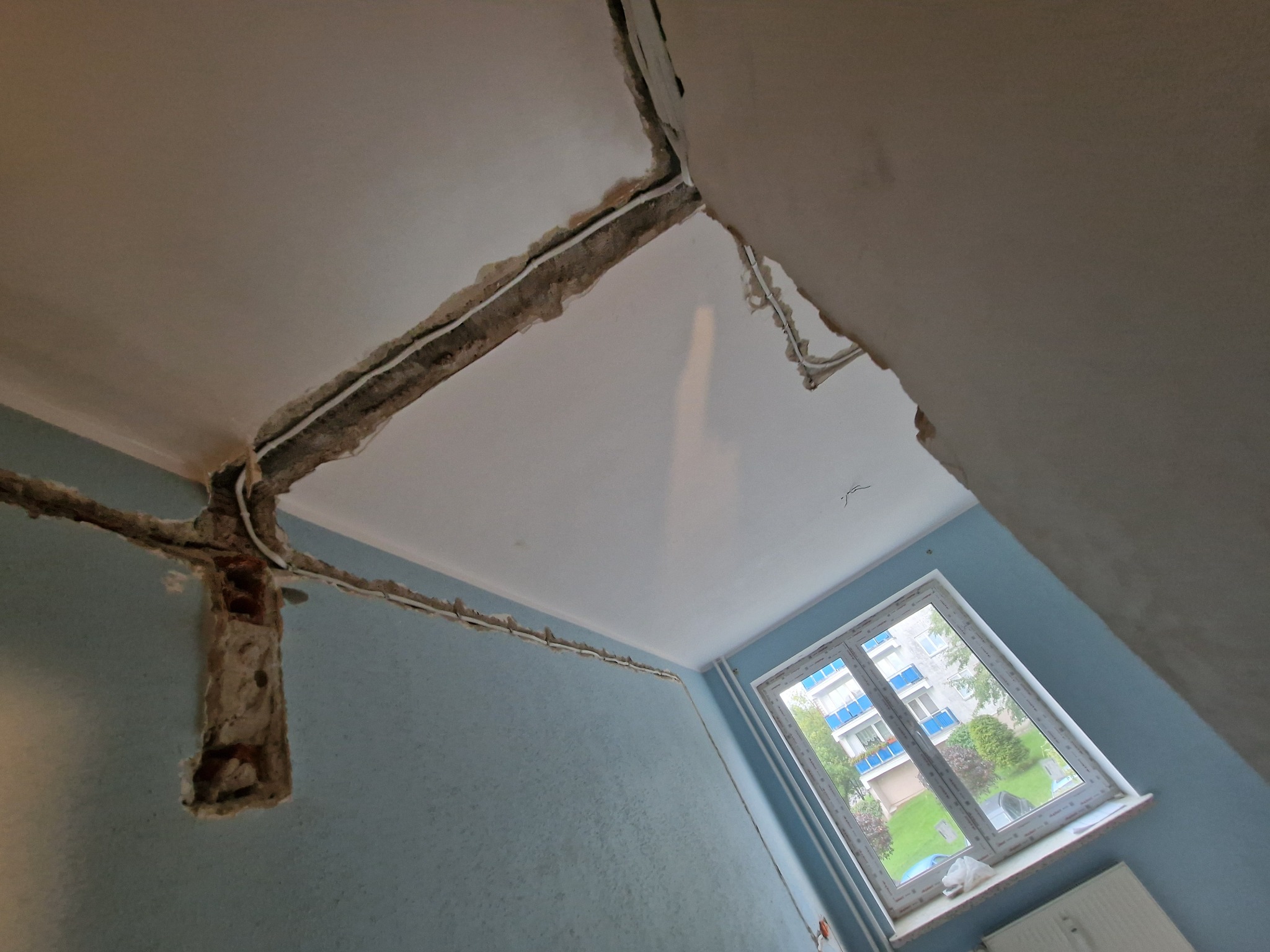 Ściana w trakcie remontu z widocznymi przewodami elektrycznymi w bruzdach, przygotowana do tynkowania. Okno z widokiem na zieleń w tle.