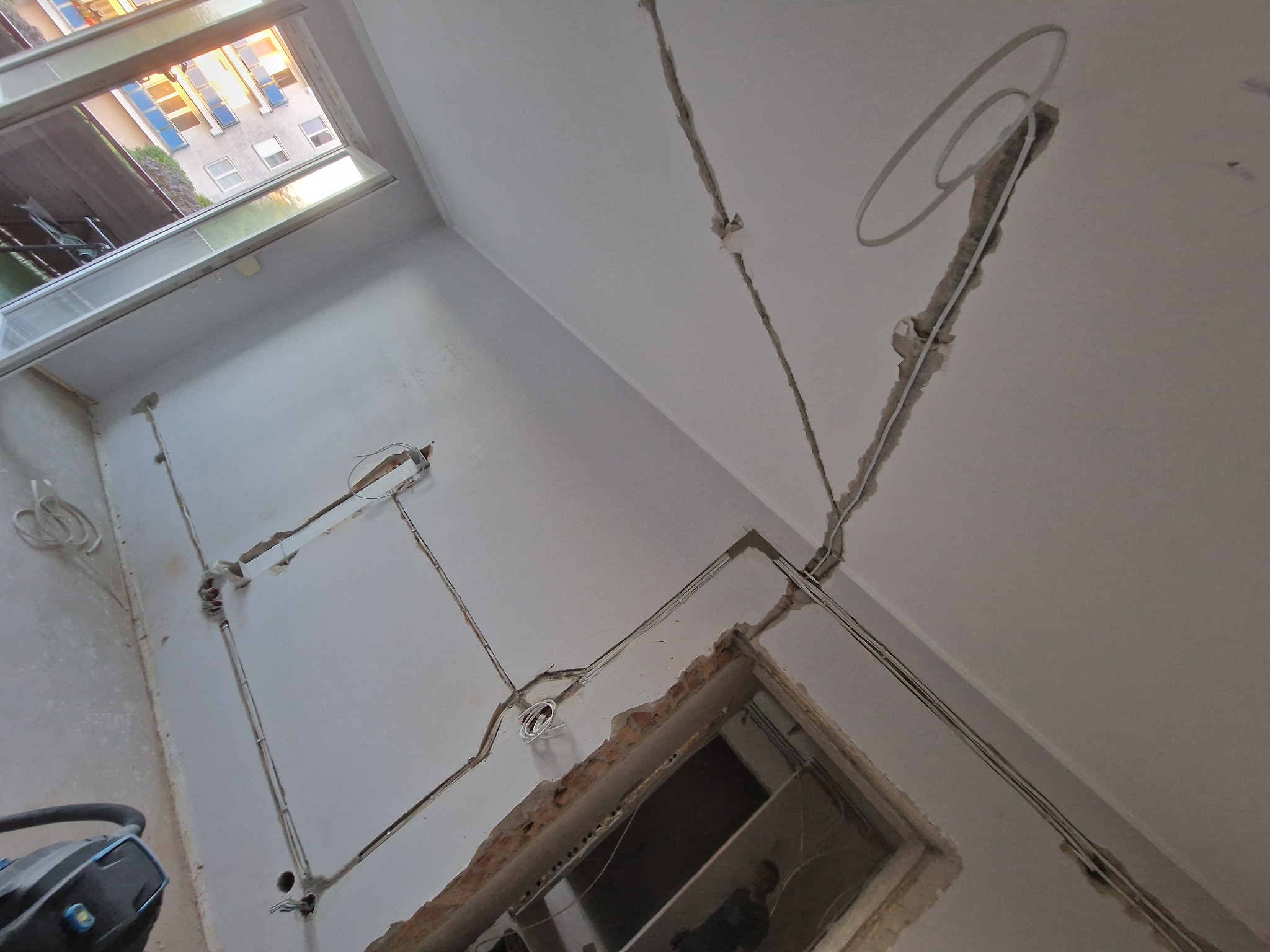 Wnętrze w trakcie remontu z widocznymi kablami elektrycznymi prowadzonymi w bruzdach w ścianie i suficie. Widoczny otwór drzwiowy i okno z widokiem na budynek.