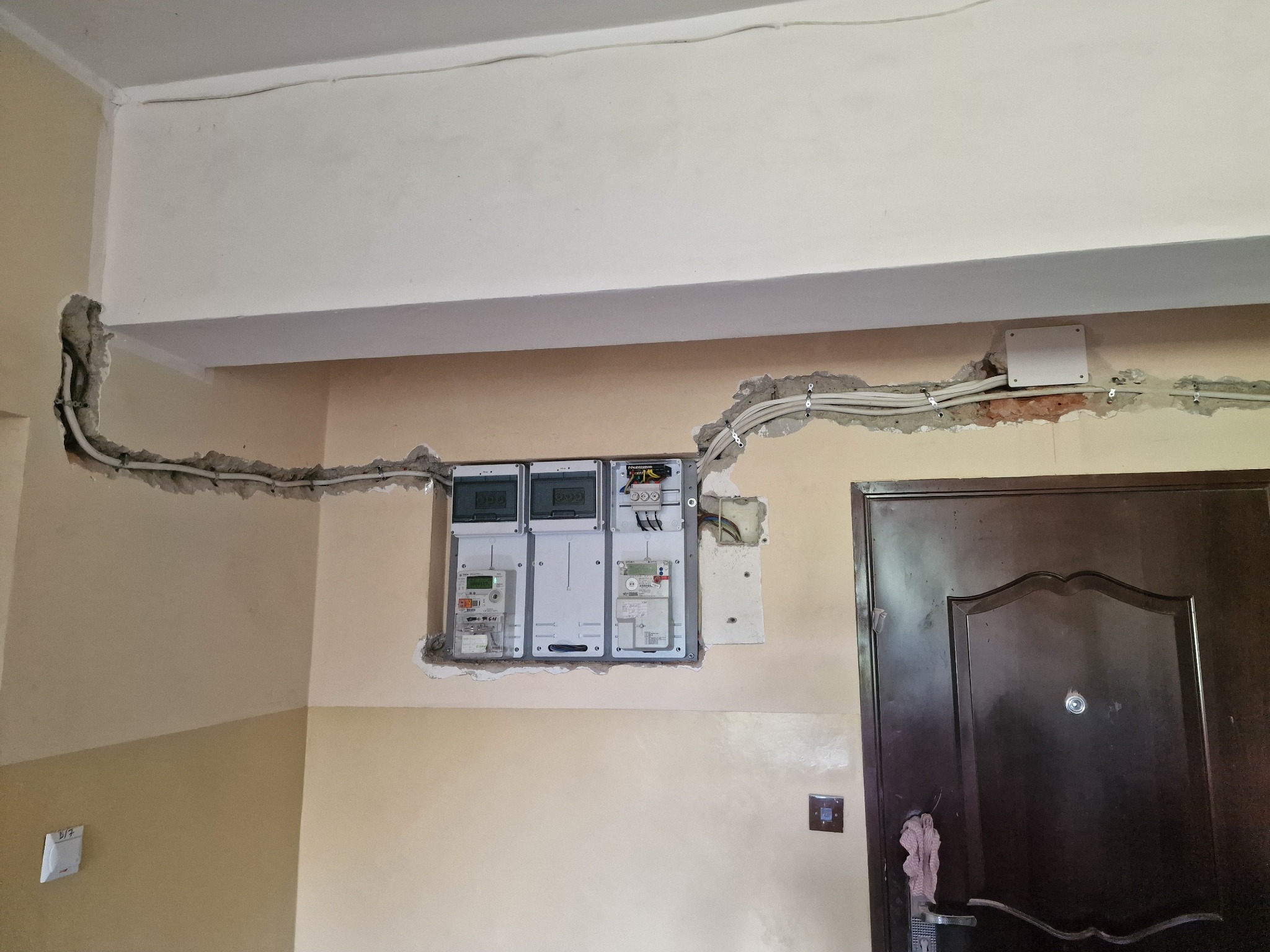 Montaż elektryki w mieszkaniu: prowadzenie kabli w bruzdach, zamontowane liczniki i skrzynka rozdzielcza, widoczne niedoskonałości ścian przed wykończeniem.