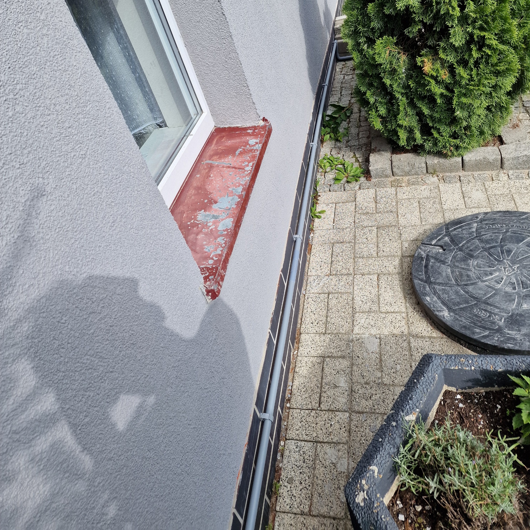 Szara ściana budynku z oknem i rurą spustową, widok z góry, kostka brukowa i rośliny. Rynna przymocowana do ściany budynku.