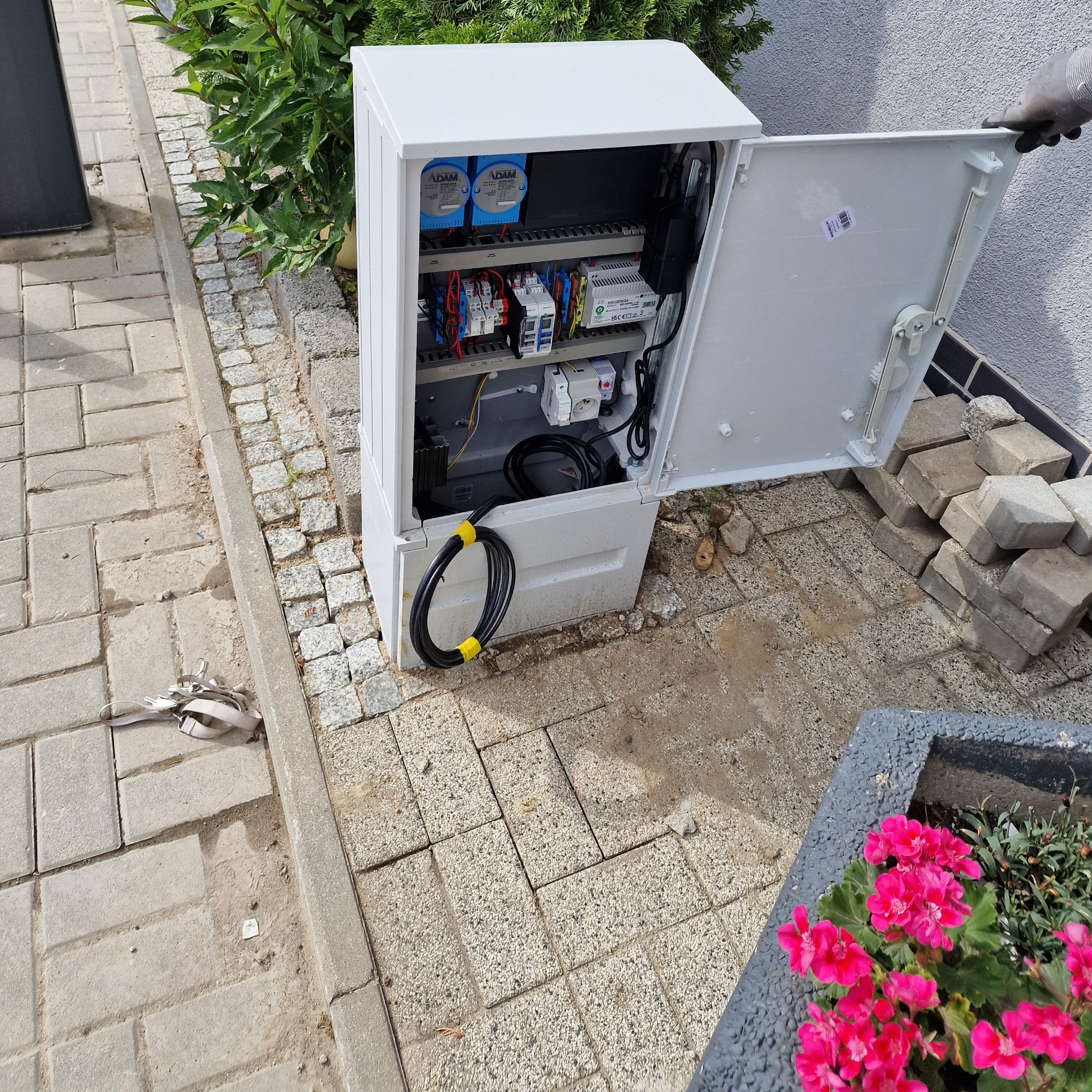 Otwarta, biała szafka elektryczna na zewnątrz budynku, z widocznymi elementami instalacji i kablem wychodzącym na chodnik z kostki brukowej, obok roślin.