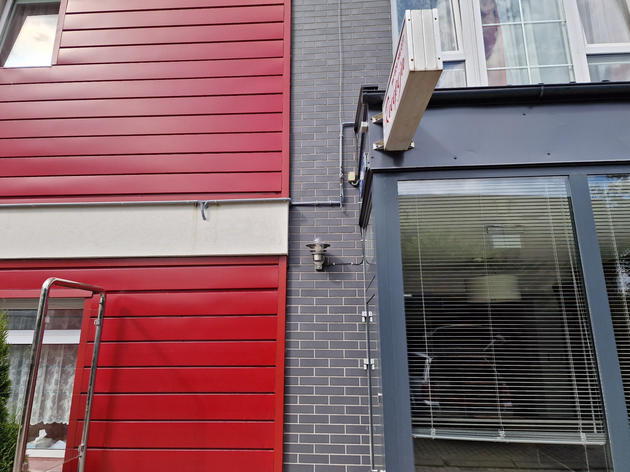 Zewnętrzna lampa ścienna na szarej, ceglanej ścianie, z widocznymi przewodami elektrycznymi i częścią czerwonej elewacji budynku. Oświetlenie zewnętrzne.