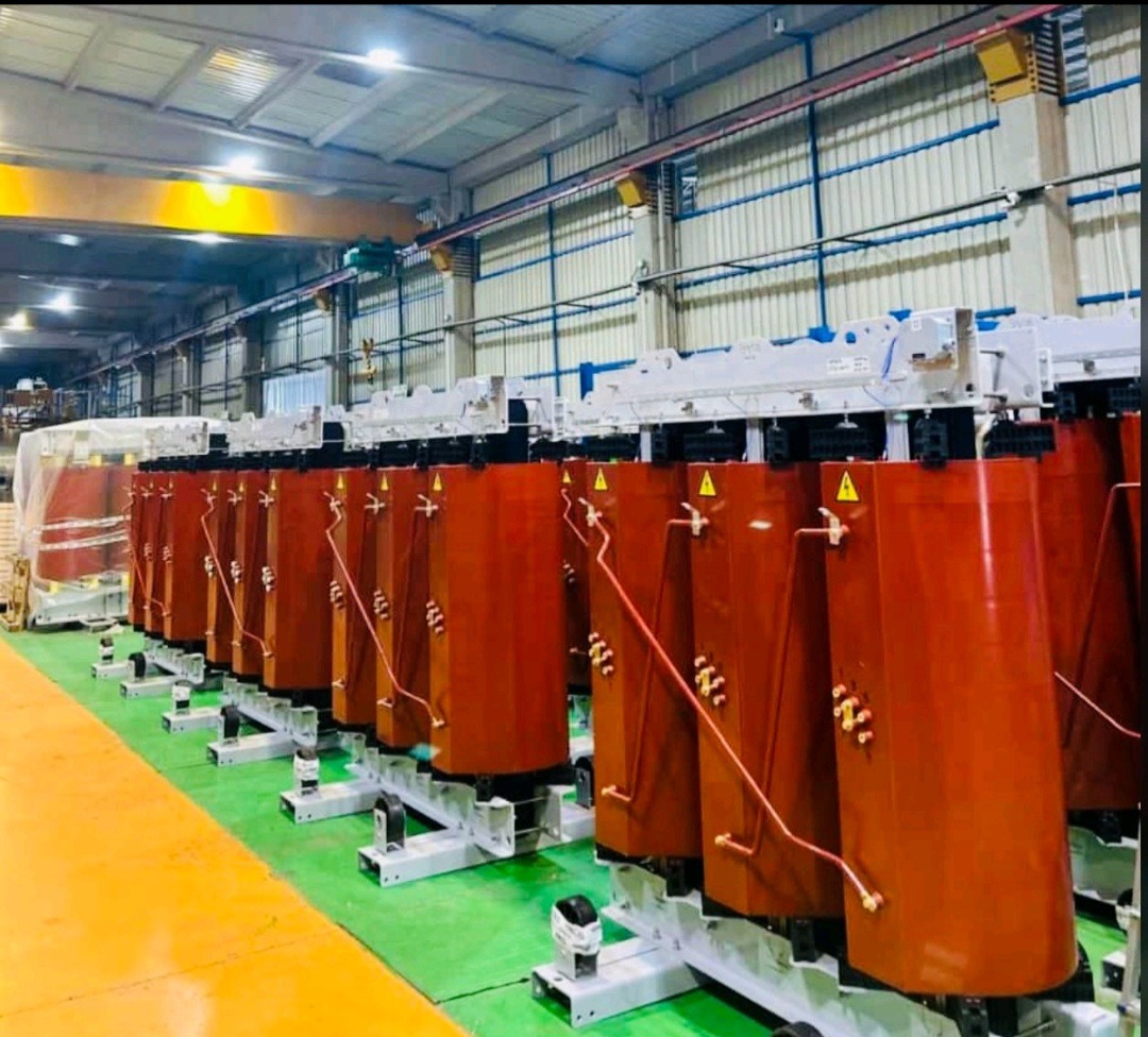 Rząd brązowych transformatorów w hali produkcyjnej, widoczne elementy instalacji i konstrukcji wsporcze. Żółty suwnicowy dźwig w tle.