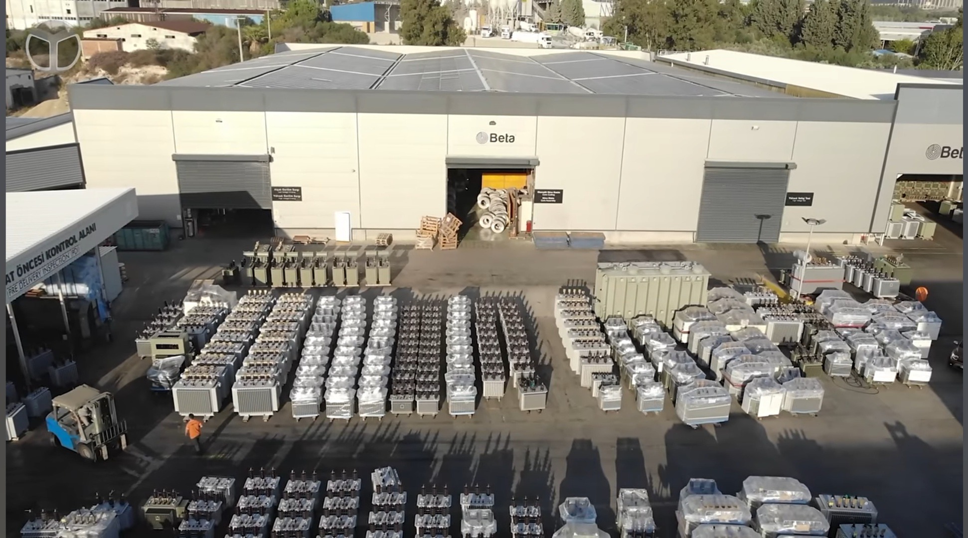 Szeroki kadr z lotu ptaka na magazyn z panelami słonecznymi na dachu. Na placu przed budynkiem zgromadzone transformatory, pracownik w pomarańczowej koszulce i wózek widłowy.