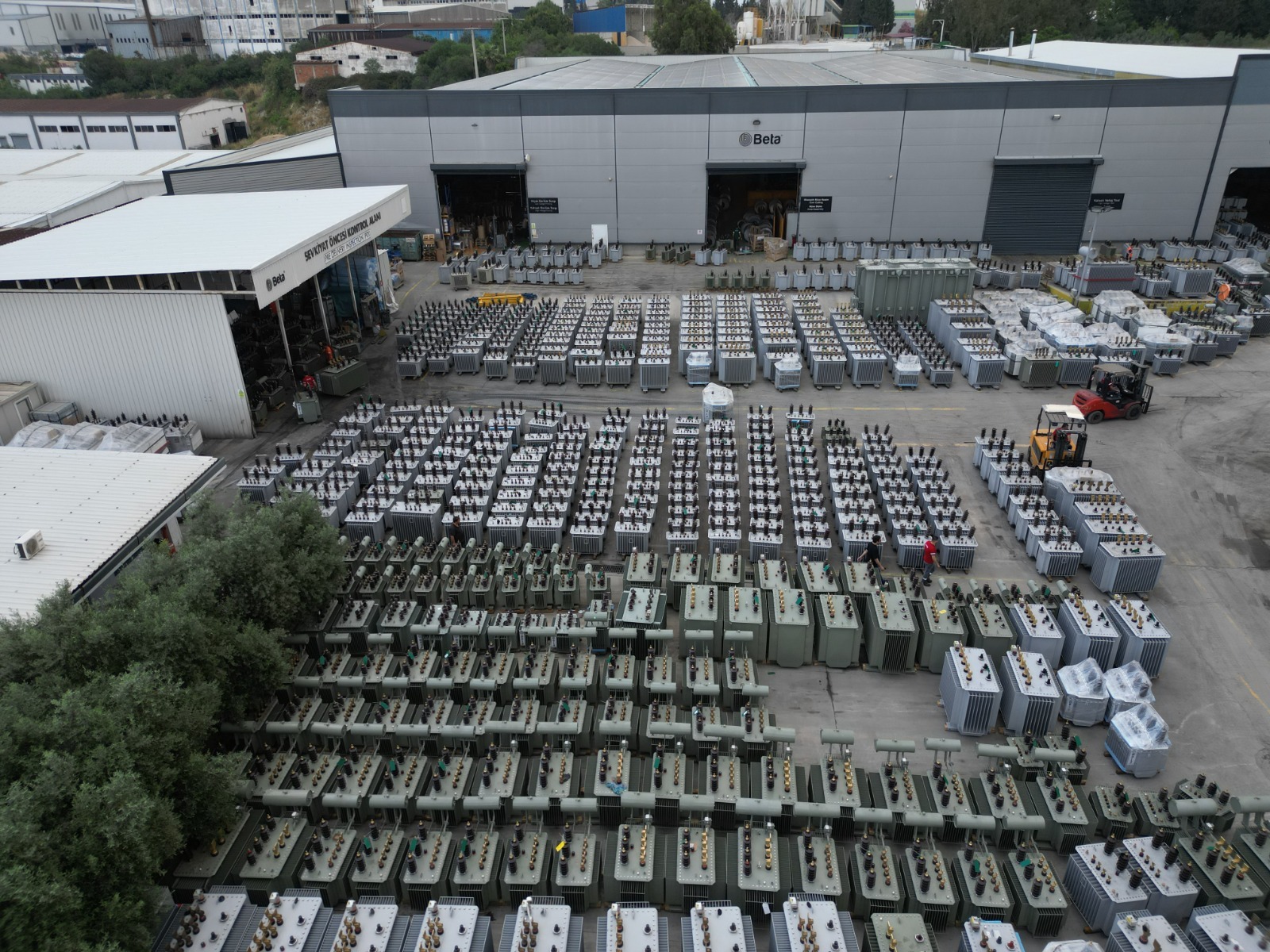Widok z góry na dziesiątki transformatorów ustawionych rzędami na placu fabryki Beta, z wózkiem widłowym i pracownikiem w czerwonym kombinezonie.