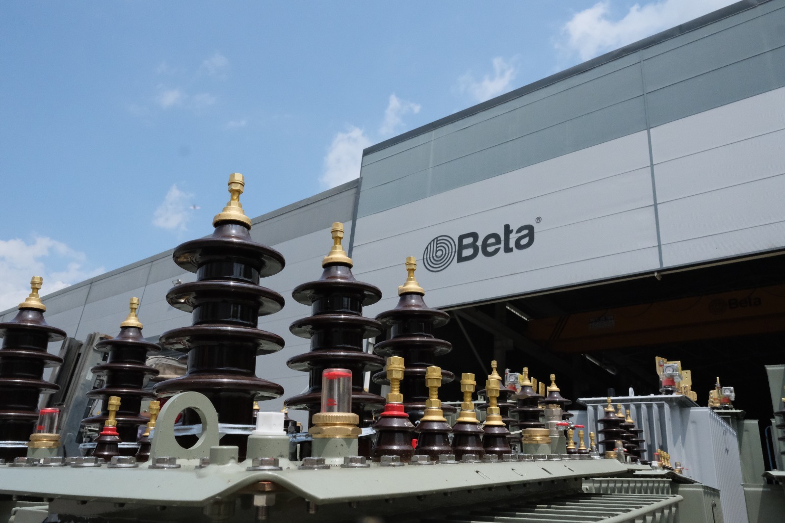 Transformator z ceramicznymi izolatorami na tle budynku z logo Beta, częściowo widoczny suwnica wewnątrz. Element instalacji elektrycznej.