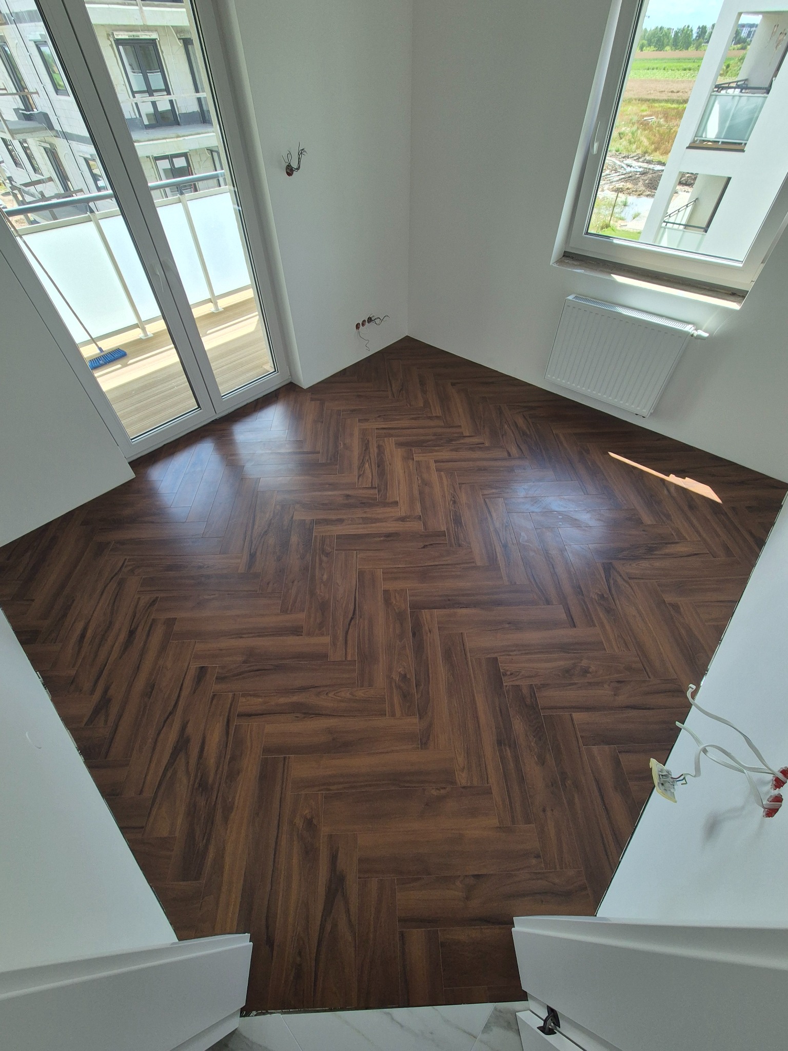 Ułożony parkiet w jodełkę w jasnym pokoju z oknami. Widoczne fragmenty balkonu i grzejnika. Perspektywa z góry.