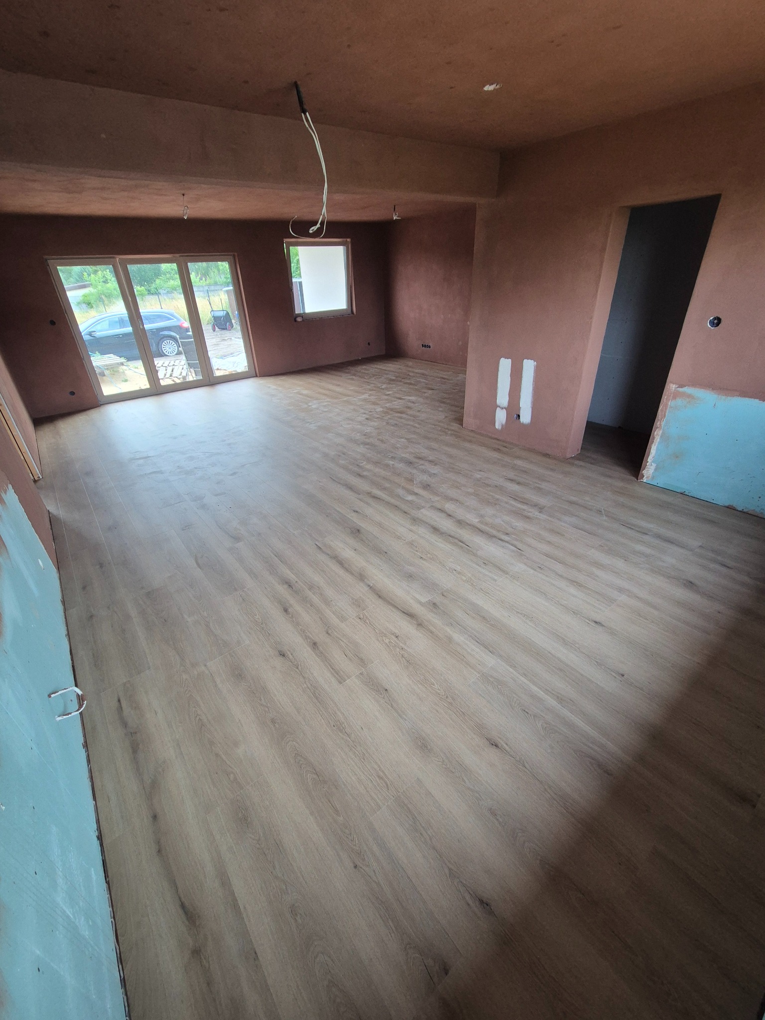 Jasne panele podłogowe ułożone w surowym wnętrzu z oknami i ścianami w odcieniach brązu, widoczne kable zwisające z sufitu. Praca w toku.