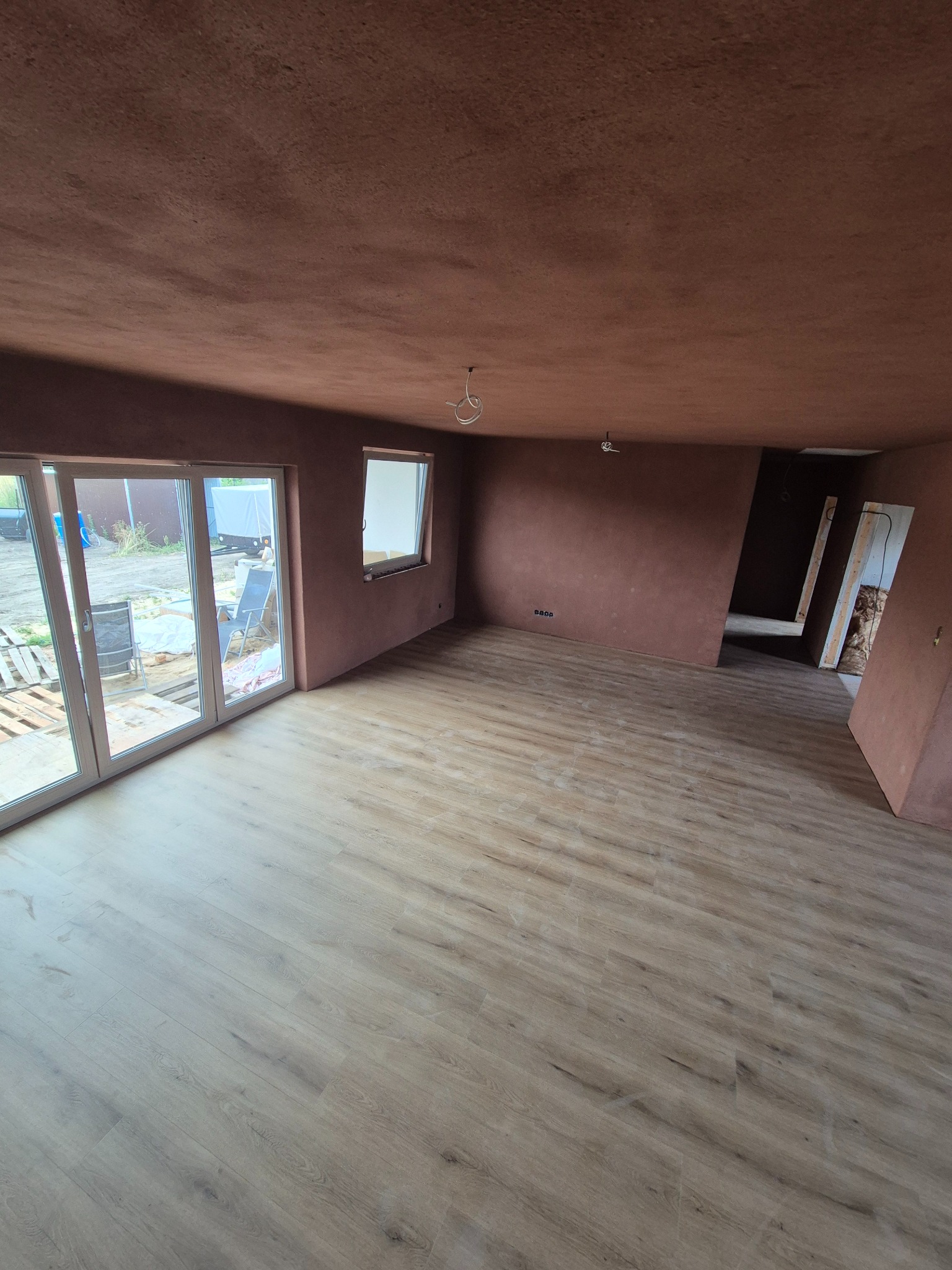 Jasne panele podłogowe w przestronnym pokoju z surowymi ścianami w odcieniach terakoty, duże okno balkonowe po lewej. Widoczne przygotowania do montażu drzwi.