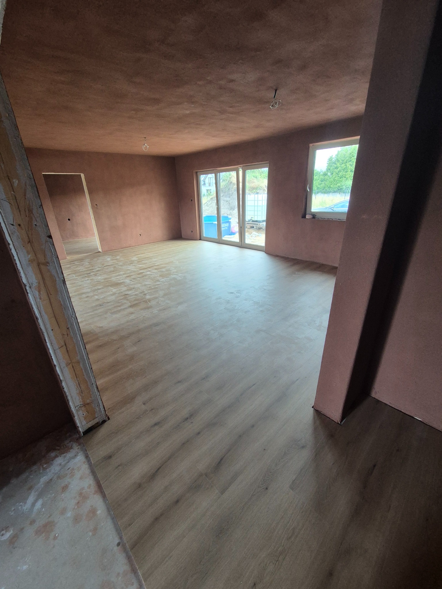 Wnętrze po remoncie: ułożony parkiet z widocznymi śladami po pracach wykończeniowych, ściany w kolorze terakoty, widoczne drzwi balkonowe i okno z widokiem na zieleń.