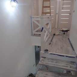 Mykola Iskovych - Białe schody i ściany w trakcie malowania, zabezpieczone folią i taśmą malarską. Widoczna drabina i akcesoria malarskie, lampa na ścianie.
