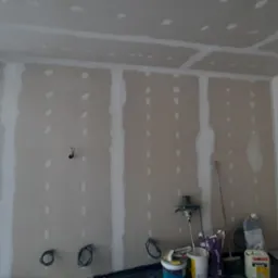 Ściana z płyt karton-gips, widoczne łączenia i otwory po wkrętach, przygotowana do szpachlowania. W tle narzędzia i materiały budowlane.