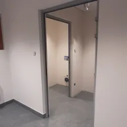 Otwarta, szara futryna drzwiowa w białym pomieszczeniu z szarą podłogą. W głębi widoczna toaleta z umywalką i instalacją.