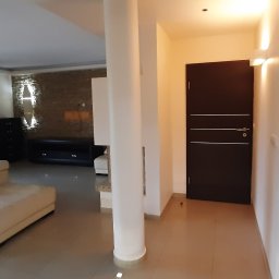 Mykola Iskovych - Jasne wnętrze domu z beżową sofą, kolumnami i dekoracyjną ścianą z kamienia. Widoczna ciemna szafka i drzwi. Oświetlenie punktowe.