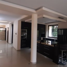 Mykola Iskovych - Nowoczesna kuchnia z wyspą i barkiem, czarne meble, jasne ściany, kolumna podtrzymująca sufit, widok na salon z dekoracyjną ścianą z kamienia.