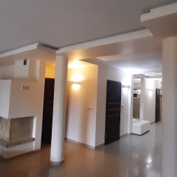 Mykola Iskovych - Przestronne wnętrze salonu z kominkiem i elementami dekoracyjnymi, utrzymane w jasnej kolorystyce z akcentami kamienia i drewna. Widoczne kolumny i nowoczesne oświetlenie.