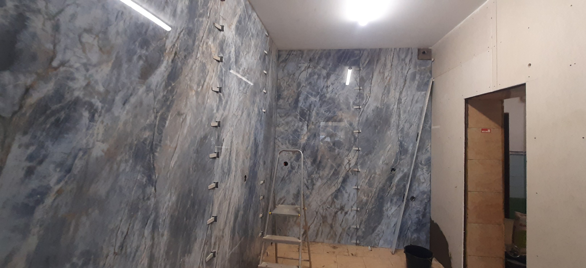 Montaż płytek imitujących marmur na ścianie, z użyciem poziomujących klipsów. Widoczna drabina i częściowo zamontowana płyta gipsowa na sąsiedniej ścianie.