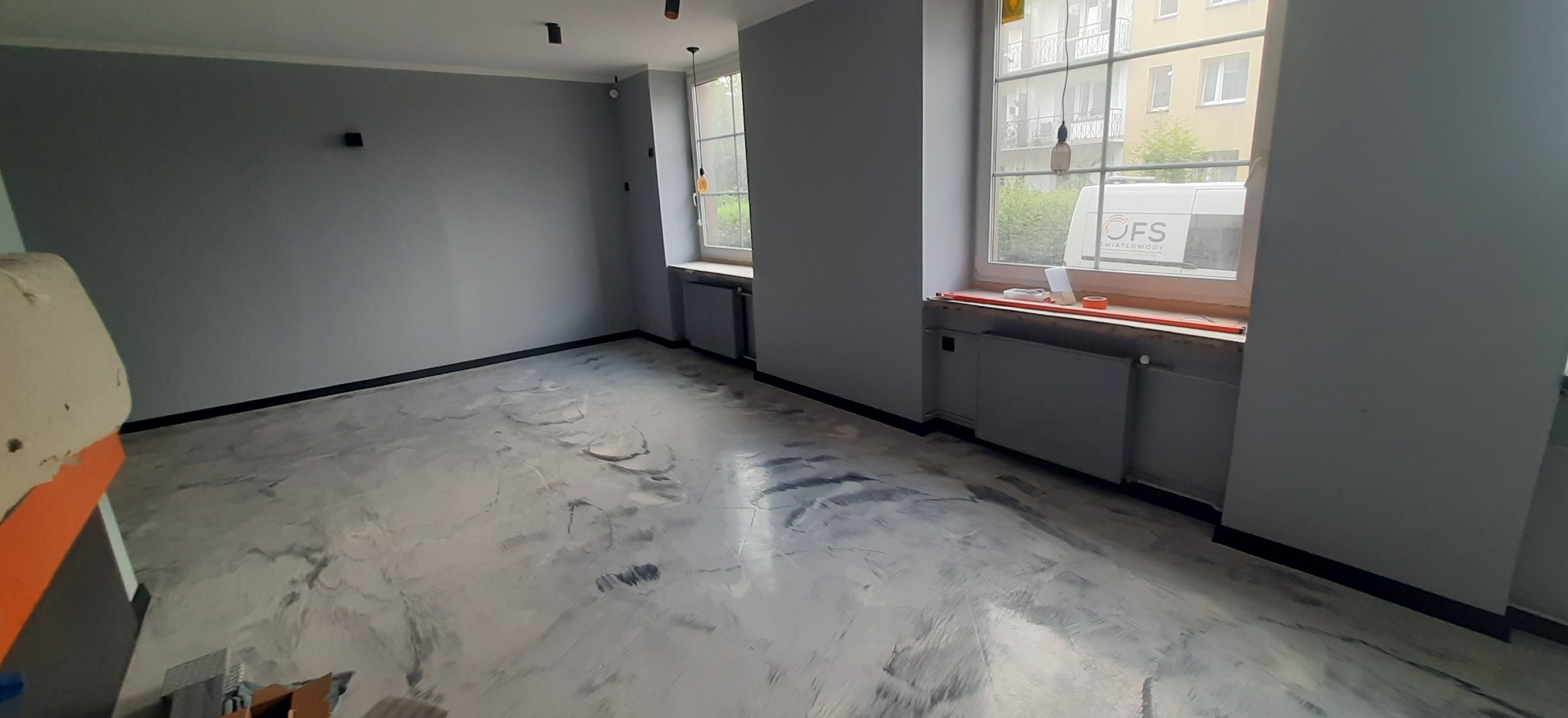 Szare, odnowione wnętrze z podłogą imitującą marmur, czarne listwy przypodłogowe i okna z widokiem na budynek. Minimalistyczne lampy sufitowe.