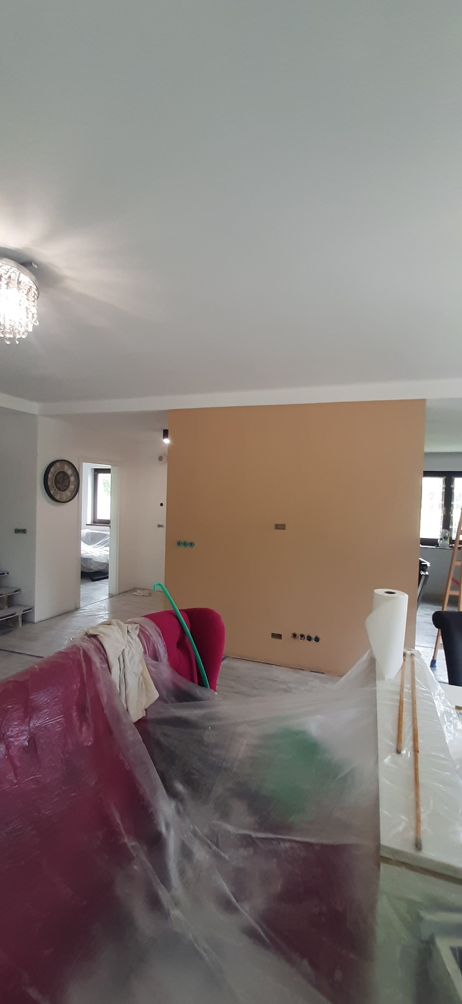 Przygotowania do malowania ścian w Jankowicach. Zabezpieczone meble folią malarską, ściana w kolorze beżowym, widoczne gniazdka elektryczne i listwy przypodłogowe.