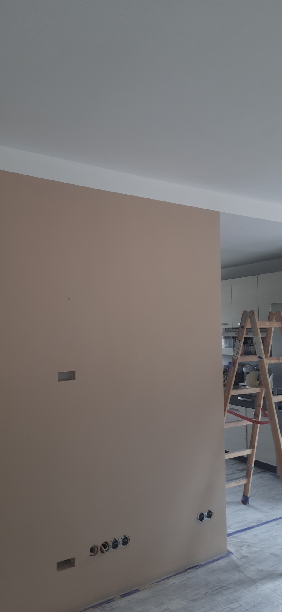 Ściana w trakcie malowania na beżowo, widoczne gniazdka elektryczne i drabina, podłoga zabezpieczona folią malarską. W tle zabudowa kuchenna.