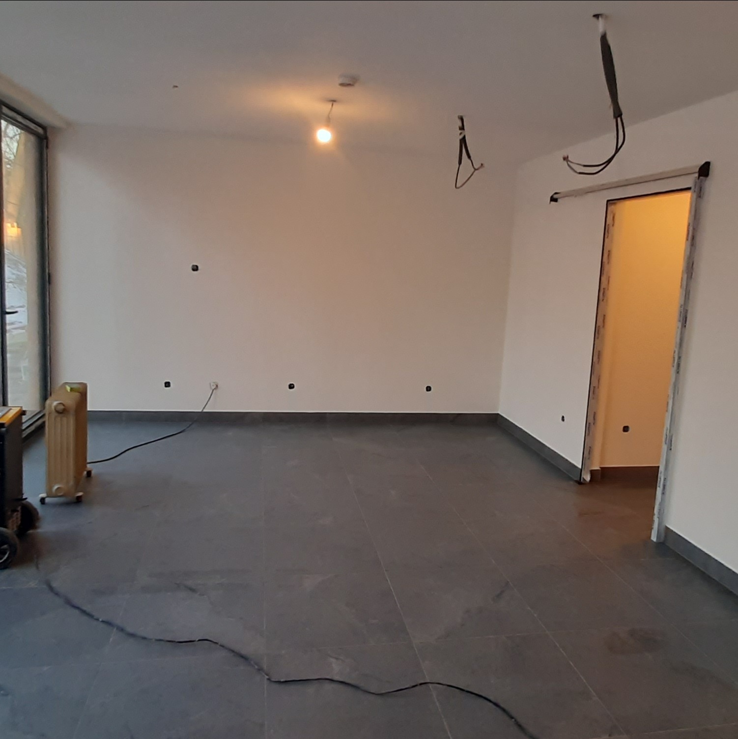 Wnętrze pokoju z szarą podłogą, białymi ścianami, oknem i otwartymi drzwiami. Widoczne niedokończone instalacje elektryczne i grzejnik olejowy.