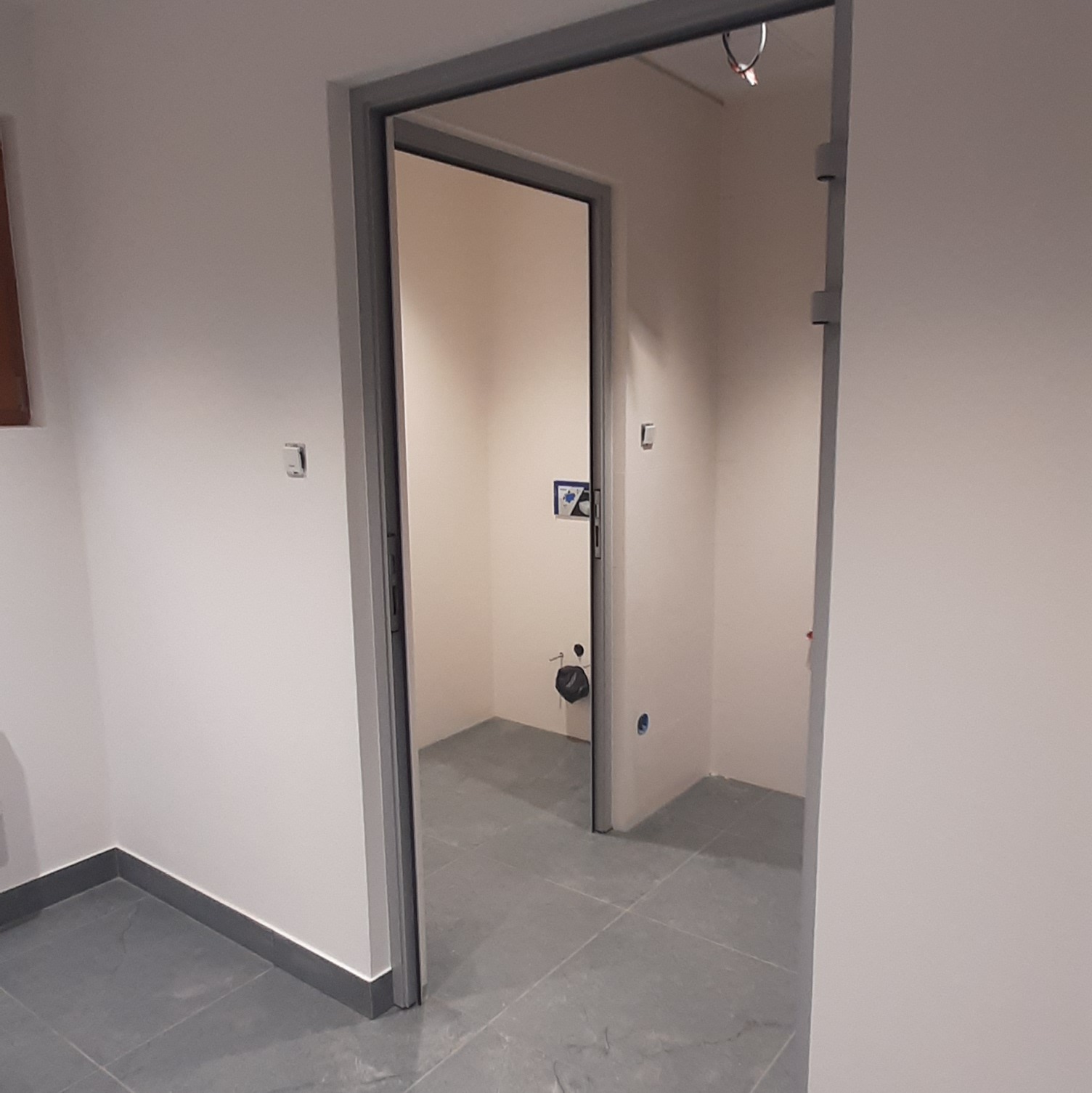 Otwarta, szara futryna drzwiowa w białym pomieszczeniu z szarą podłogą. W głębi widoczna toaleta z umywalką i instalacją.