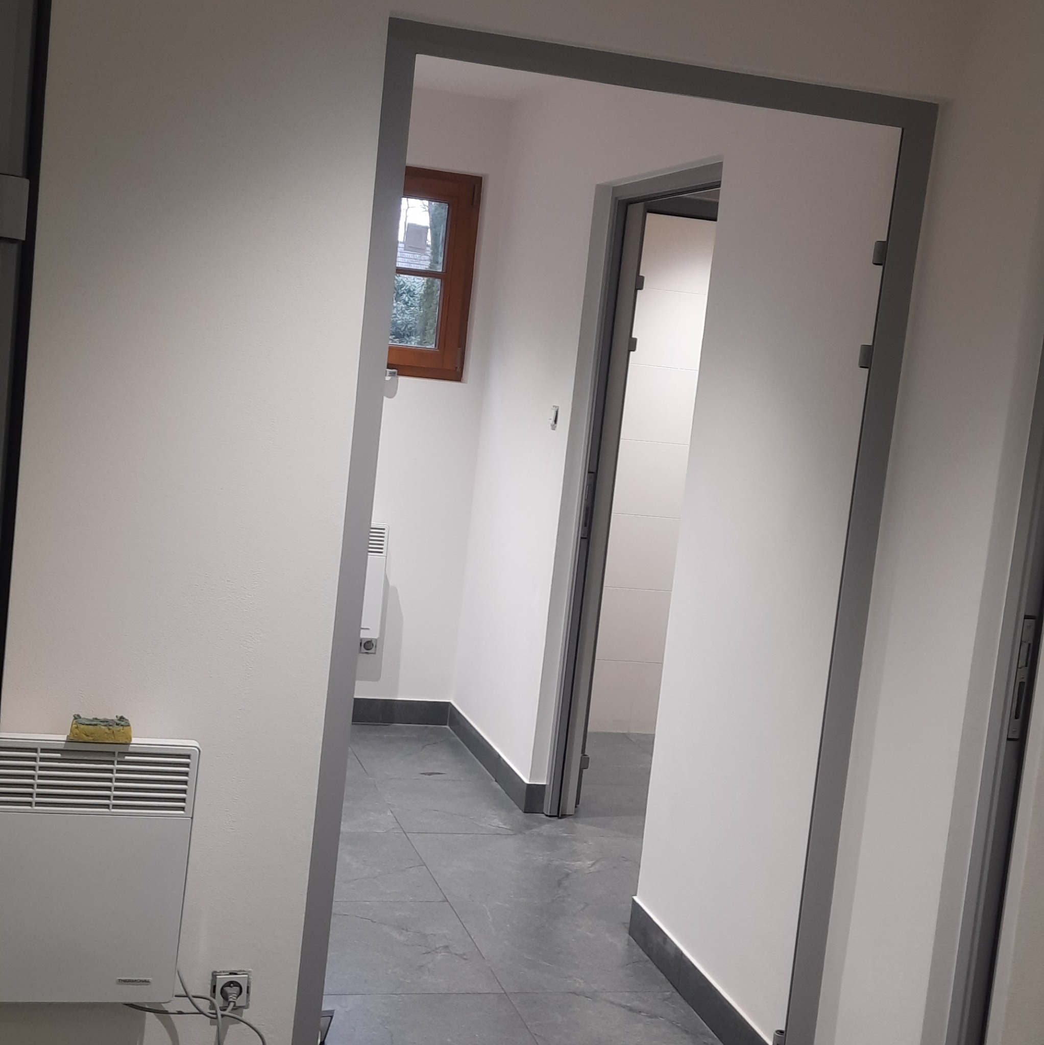 Wykończone wnętrze łazienki z szarą podłogą, widok przez dwa otwory drzwiowe z szarymi ościeżnicami. Białe ściany, grzejnik i okno w głębi.