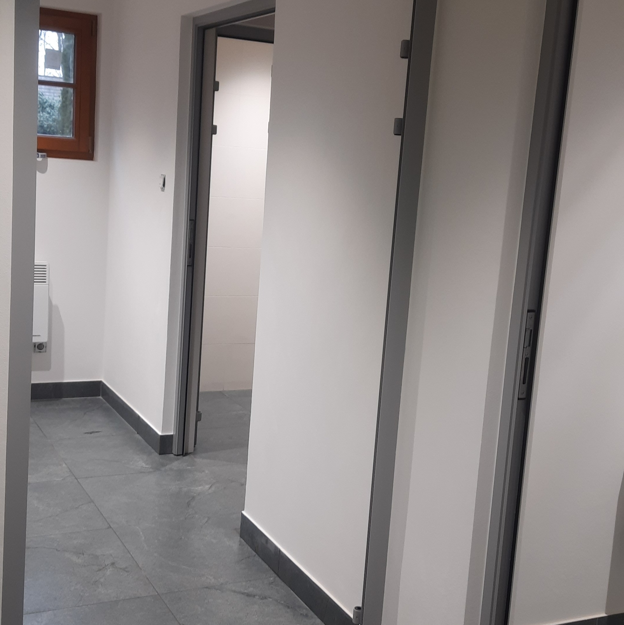 Nowoczesne wnętrze łazienki z szarą podłogą i białymi ścianami, widoczne dwie otwarte kabiny z szarymi futrynami drzwi. Minimalistyczny design z oknem w tle.