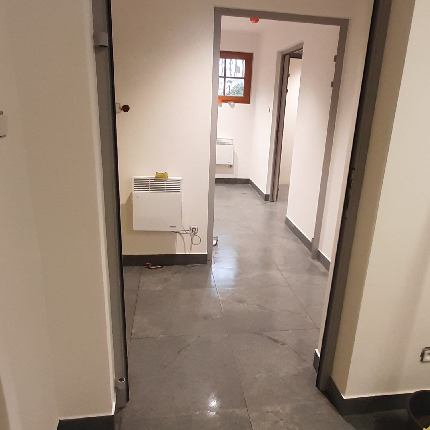 Korytarz po remoncie z szarą podłogą, świeżo pomalowanymi ścianami i nowymi drzwiami. Widoczny grzejnik na ścianie i okno w głębi.