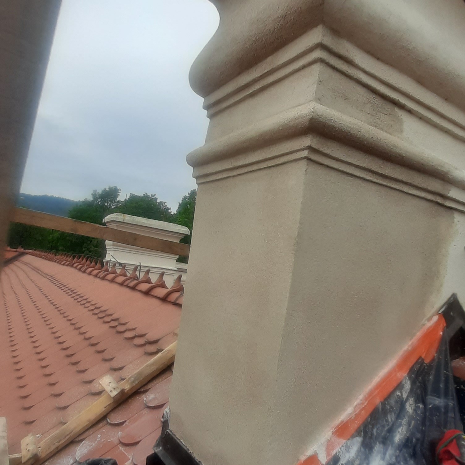 Wykończona, jasna kolumna architektoniczna z detalami, widoczna na tle czerwonej dachówki i zieleni. Element dekoracyjny elewacji po renowacji.