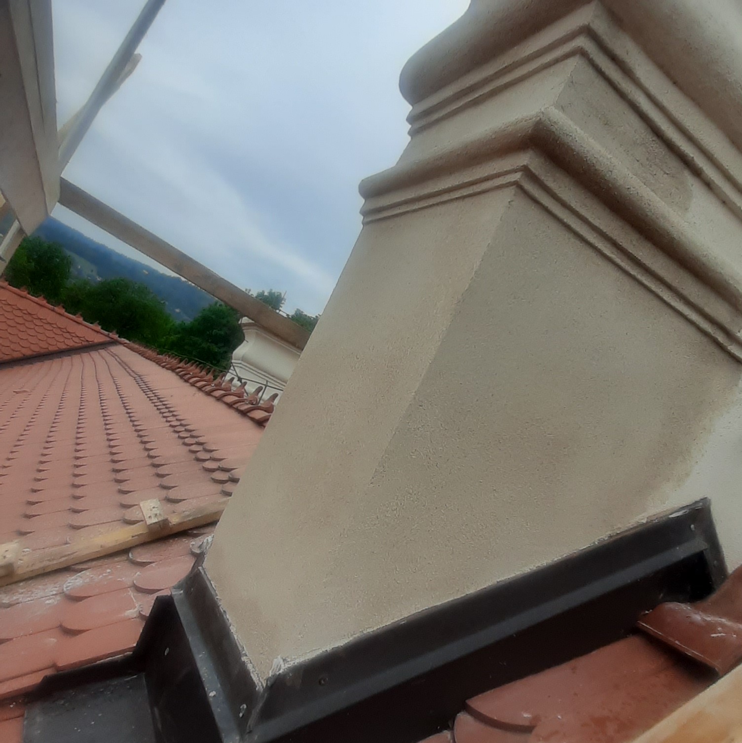 Otynkowany komin na dachu z czerwoną dachówką, widok z bliska. Fragment rusztowania w tle. Detal architektoniczny po renowacji.