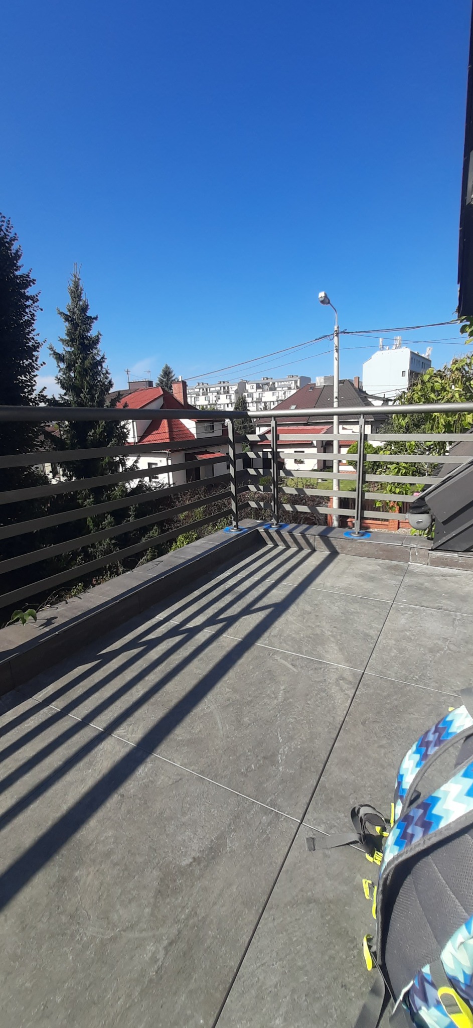 Szare płytki na balkonie w słoneczny dzień, widok na budynki i drzewa przez metalową barierkę. Cienie tworzą geometryczny wzór na podłodze. Fragment plecaka w rogu.