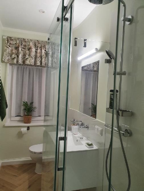 Jasna łazienka po remoncie: kabina prysznicowa ze szklanymi drzwiami, umywalka z szafką, toaleta i okno z zasłonką w liście. Parkiet ułożony w jodełkę.