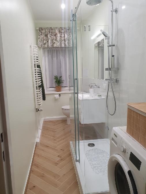 Jasna łazienka po remoncie: kabina prysznicowa ze szklanymi drzwiami, drewnopodobna podłoga w jodełkę, biała umywalka i pralka. Roślina w oknie.