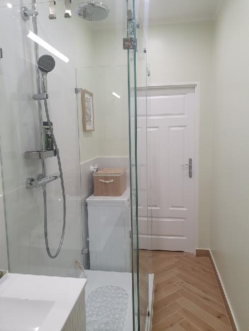 Nowoczesna łazienka z kabiną prysznicową typu walk-in, pralką ukrytą pod blatem i drewnianą podłogą ułożoną w jodełkę. Minimalistyczny design i jasna kolorystyka.