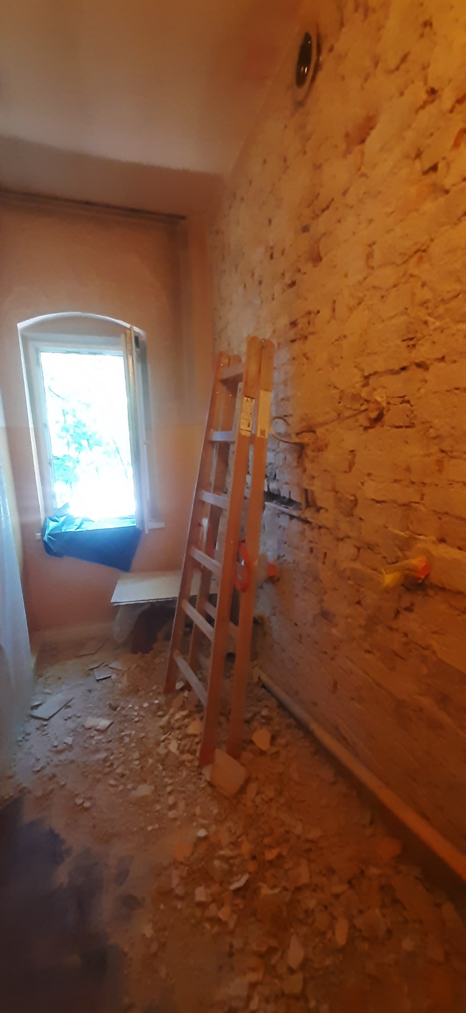 Wnętrze w trakcie remontu: odsłonięta cegła, drabina oparta o ścianę, gruz na podłodze. Widoczne instalacje elektryczne i okno z niebieską zasłoną.